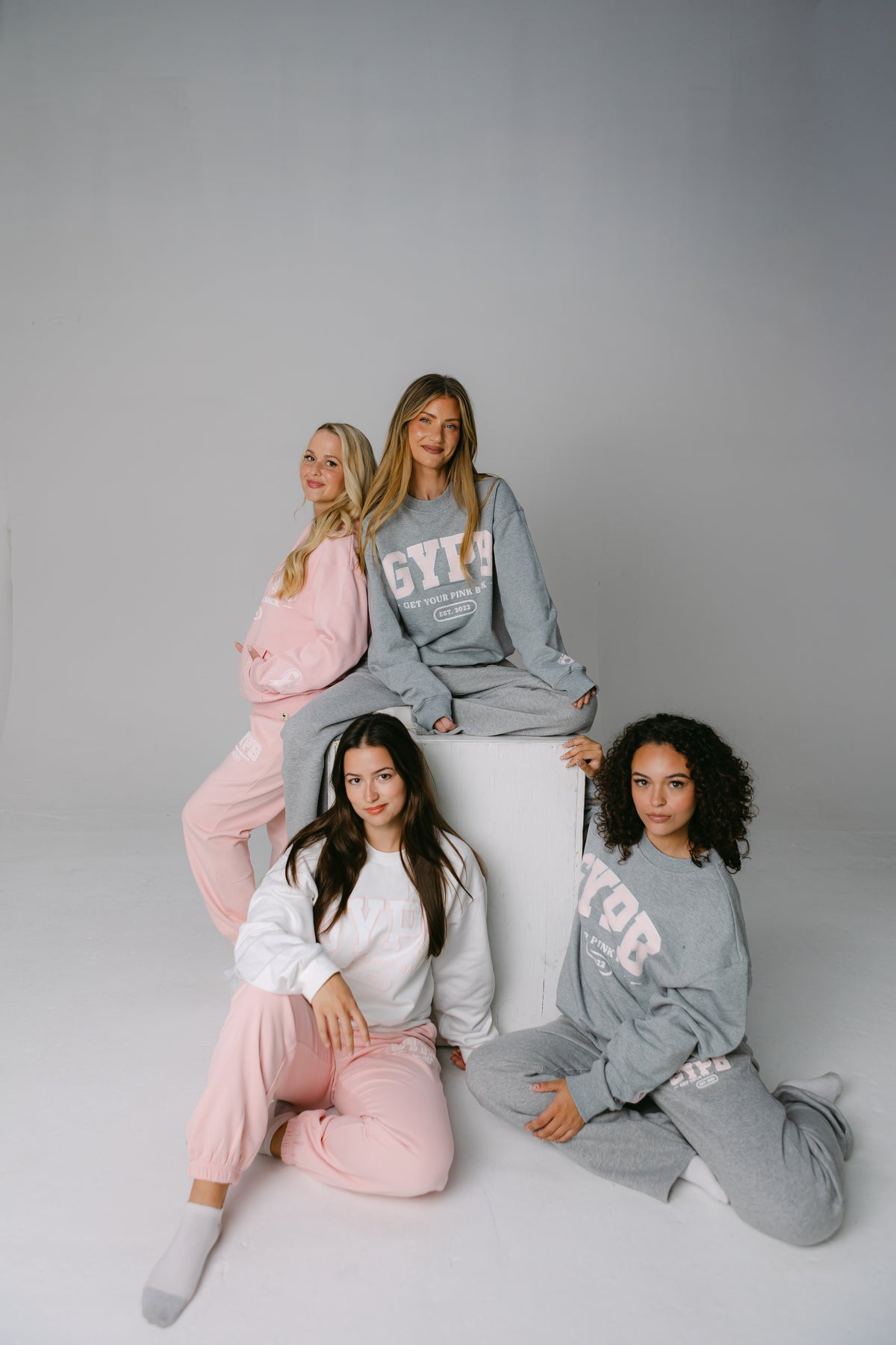 Four women in Get Your Pink Back crewnecks, hoodies and joggers posed around a white block